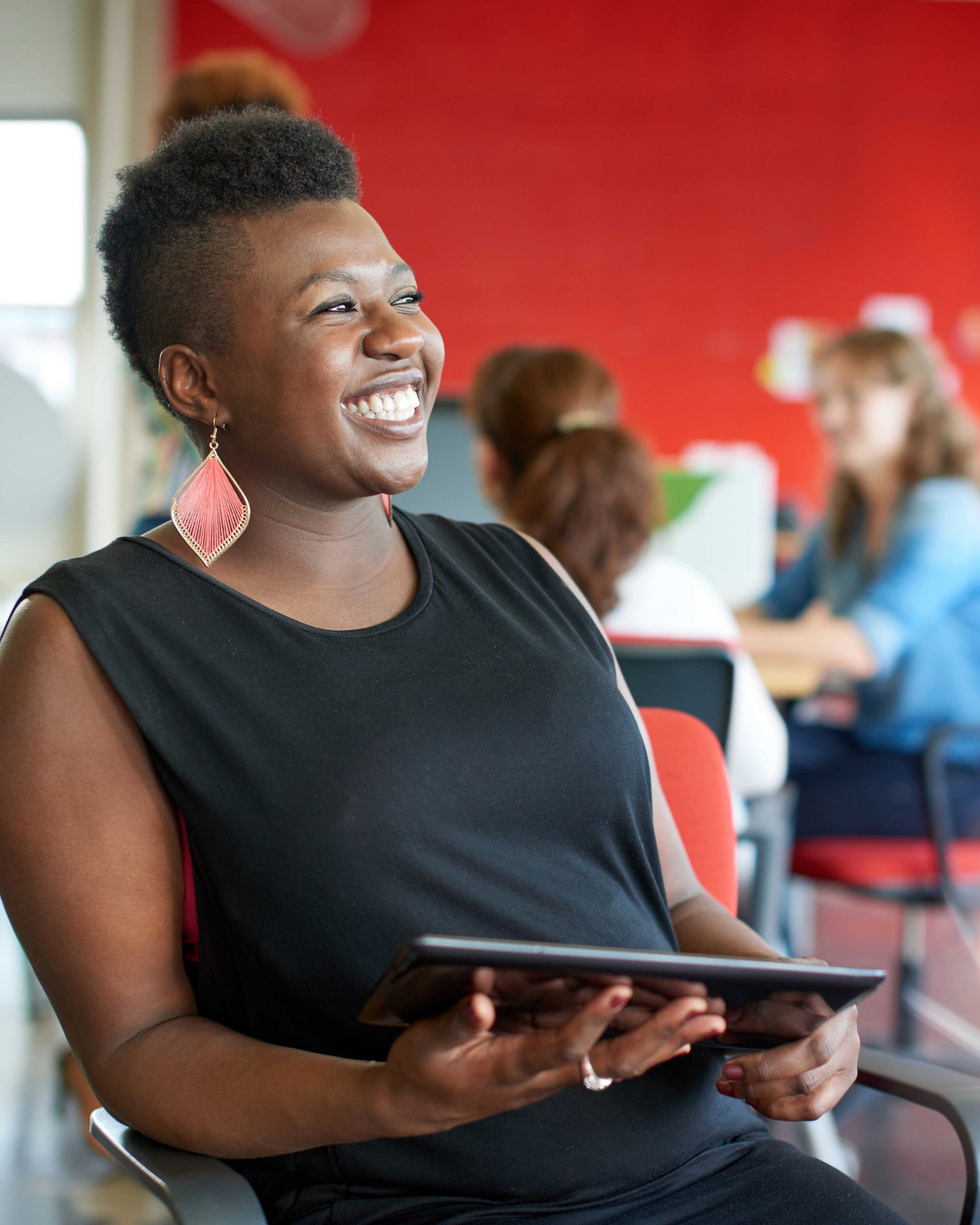 A woman with a short hairstyle and large earrings smiles while holding a tablet in a bright office setting with colleagues blurred in the background.