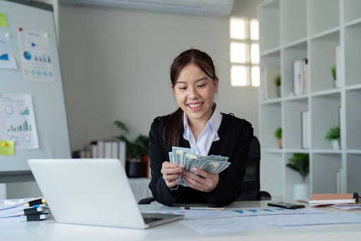 Smiling businesswoman counting money at her office desk with laptop and financial documents.