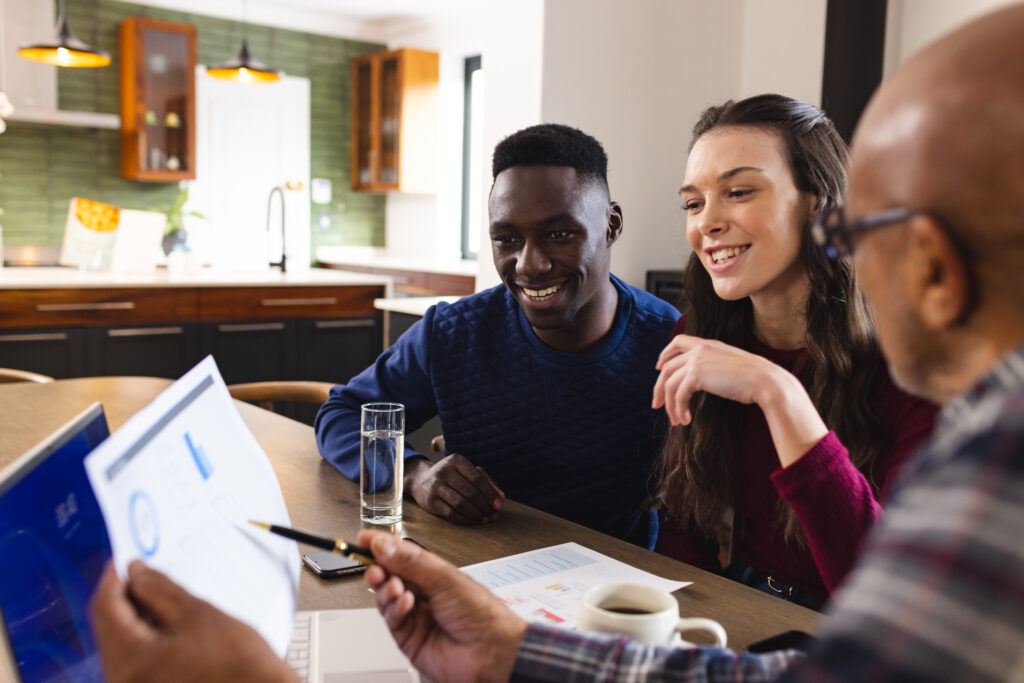 Diverse couple and biracial male financial advisor using laptop and discussing documents at home. Finance, communication, home budget and financial advice, unaltered.